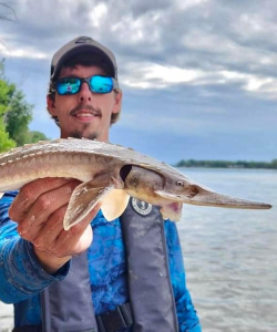 Stefan R. Tucker holding a small fish in the foreground of the image with rippled water in the background.